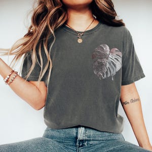 May include: Dark gray t-shirt featuring a large, stylized leaf design on the front. The leaf is a mix of silver and gray tones. The shirt has a relaxed fit and is paired with blue jeans.
