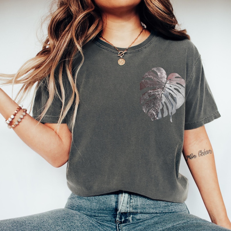 May include: Dark gray t-shirt featuring a large, stylized leaf design on the front. The leaf is a mix of silver and gray tones. The shirt has a relaxed fit and is paired with blue jeans.