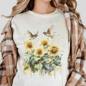 Op de afbeelding: Crèmekleurig T-shirt met een aquarelontwerp van zonnebloemen en twee vogels in vlucht. De zonnebloemen hebben gele bloemblaadjes en bruine centra, met groene bladeren en stelen. De vogels zijn bruin en wit. Gedragen met een bruin geruit overhemd.