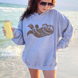 May include: A gray hooded sweatshirt with a brown graphic of two otters cuddling. The sweatshirt is worn by a person standing on a beach.