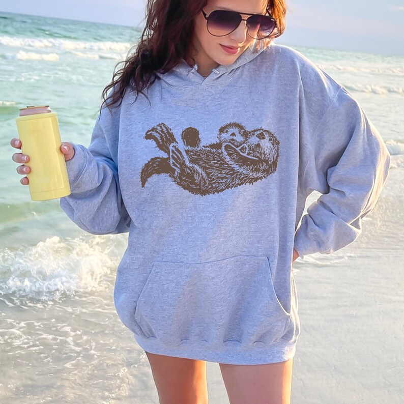 May include: A gray hooded sweatshirt with a brown graphic of two otters cuddling. The sweatshirt is worn by a person standing on a beach.