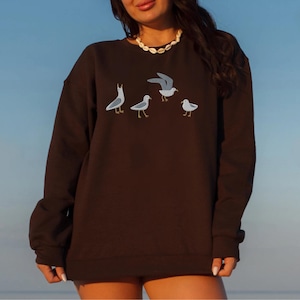 May include: A dark brown sweatshirt featuring an embroidered design of four seagulls in various poses. The birds are depicted in shades of gray and white with yellow feet. The sweatshirt has a crew neck and long sleeves.