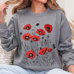 May include: A gray sweatshirt featuring a floral design of red poppies with black centers and stems. The poppy flowers are the focal point, with detailed petals and dark seed pods. The sweatshirt is a casual garment.