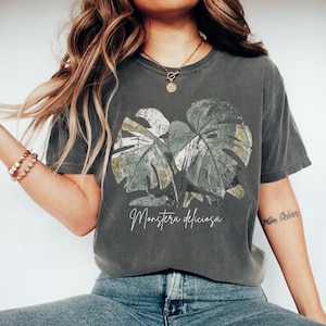 Op de afbeelding: Donkergrijs t-shirt met een botanische illustratie van Monstera deliciosa bladeren. De afbeelding is in groen, wit en bruin. De tekst "Monstera deliciosa" staat in een scriptletter onder de bladeren.