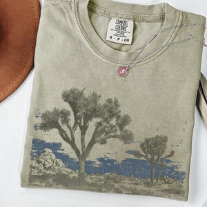 Op de afbeelding: Olijfgroen Comfort Colors T-shirt met een woestijnlandschap. Het ontwerp toont Joshua-bomen en een blauwe lucht. Een zilveren ketting met een roze hanger ligt op het shirt.