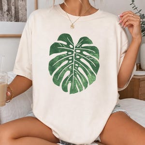 May include: An off-white short-sleeve t-shirt featuring a large, centered graphic of a green monstera leaf. The leaf design has a textured, distressed appearance. The shirt has a crew neckline and a loose, relaxed fit, providing a casual look.