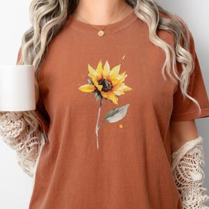 Op de afbeelding: Roestkleurig T-shirt met een aquarel zonnebloemontwerp. De zonnebloem heeft gele bloemblaadjes, een donker centrum en een groene stengel en blad. Het shirt is een korte mouw ronde hals.