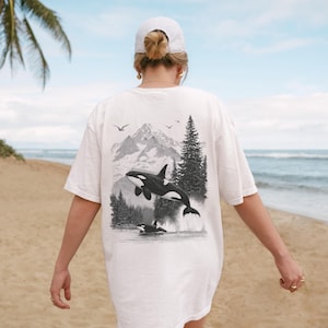 May include: White t-shirt featuring a black and white graphic of orcas in a mountain and forest landscape. The design includes a jumping orca and two others in the water, with birds flying above. The shirt is worn on a beach.