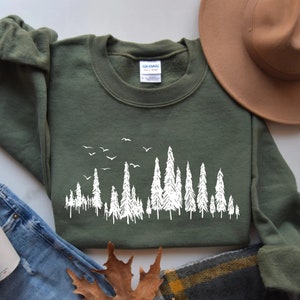 May include: Olive green sweatshirt with a white forest and birds graphic. The crew neck sweatshirt is made of a soft material. A brown hat and blue jeans are next to the sweatshirt.