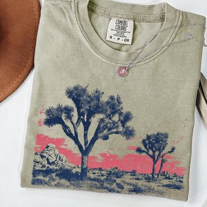 Op de afbeelding: Saliegroen t-shirt met een woestijnlandschap ontwerp. Het ontwerp toont Joshua-bomen in marineblauw en een roze lucht. Het shirt heeft een Comfort Colors label en een zilveren ketting met een hanger.