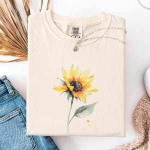 Op de afbeelding: Crèmekleurige Comfort Colors sweatshirt met een aquarel zonnebloemontwerp. De zonnebloem heeft gele bloemblaadjes en een donker centrum, met groene bladeren en stengel. Een zilveren ketting met een kleine hanger rust op de trui. De trui is op een wit oppervlak gevouwen.