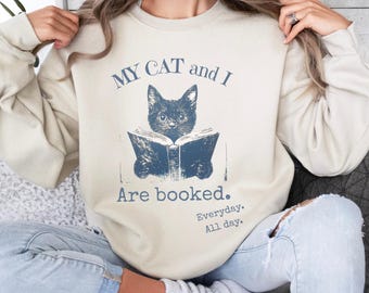 May include: Cream-colored sweatshirt with a blue graphic of a cat reading a book. The text above the cat reads "MY CAT and I", and below the cat, it says "Are booked. Everyday. All day."
