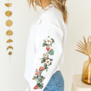 Peut inclure: Sweat-shirt blanc &agrave; manches longues avec un motif floral et des fraises. Le motif est dans les tons de rouge, vert et blanc.