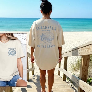 Può includere: T-shirt color crema con la scritta "SEASHELLS ARE MY HOME" e "PLEASE LEAVE THEM ON THE BEACH" in azzurro. È presente anche una grafica a conchiglia. La maglietta è indossata su una passerella sulla spiaggia con l'oceano sullo sfondo.