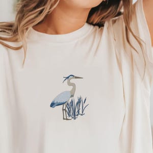 Op de afbeelding: Crèmekleurig T-shirt met een geborduurde blauwe reiger die in riet staat. De reiger heeft een lange nek en snavel, met blauwe en grijze veren. Het riet is donkerblauw-groen.