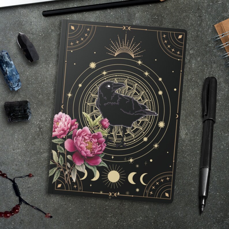 May include: Black and gold journal with a crow and floral design. The cover features a black crow with a gold sun and moon design. The background is black with gold accents.