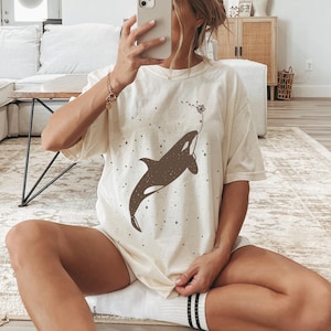 Op de afbeelding: Een wit T-shirt met een bruin silhouet van een orka die zwemt met een paardenbloem die in de wind waait. Het shirt heeft een subtiel glitterpatroon.