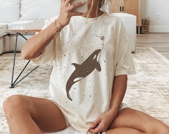 May include: A white t-shirt featuring a brown silhouette of an orca swimming with a dandelion blowing in the wind. The shirt has a subtle sparkle pattern.
