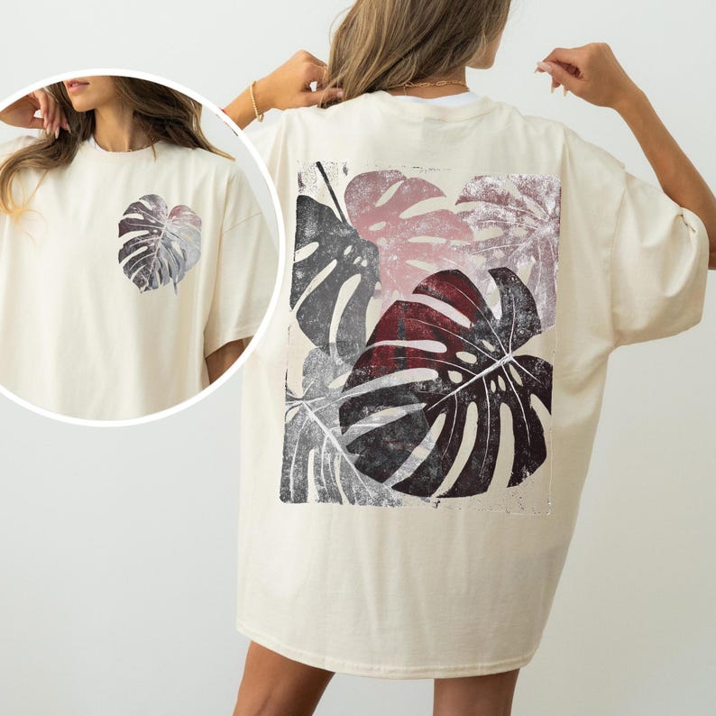 May include: Cream-colored t-shirt featuring a large, stylized monstera leaf print on the back and a smaller leaf on the front. The leaves are in shades of gray, burgundy, and pink. The shirt has a relaxed fit and short sleeves.