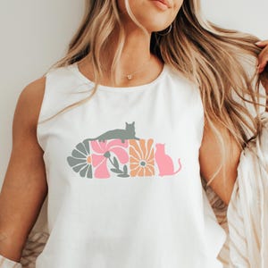 Op de afbeelding: Witte mouwloze top met een bloemen- en kattenontwerp. De afbeelding bevat een grijze kat, roze en oranje bloemen en een roze katten silhouet. Het ontwerp is gecentreerd op de voorkant van de mouwloze top.