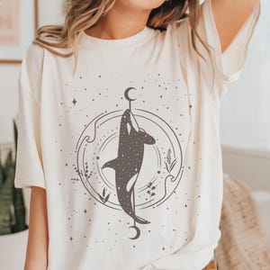 Op de afbeelding: Een wit T-shirt met een zwarte afbeelding van een orka die in een cirkel met sterren en manen zwemt. De orka wordt omringd door een cirkel met golven en bloemmotieven.