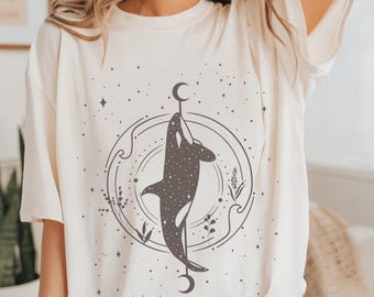 May include: A white t-shirt featuring a black graphic of an orca swimming in a circle with stars and moons. The orca is surrounded by a circle with waves and floral designs.