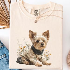 May include: Cream-colored t-shirt with a graphic of a Yorkshire Terrier wearing a floral sweater and a dark green turtleneck. The shirt is folded, with the "Comfort Colors" brand visible. A gold necklace with a small pendant is draped across the shirt.
