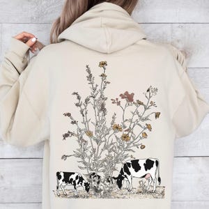 May include: Beige hoodie with a floral and cow graphic on the back. The design features a detailed illustration of wildflowers in shades of brown and yellow, with two black and white cows grazing below.