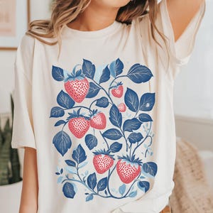 May include: A white t-shirt with a blue and red strawberry print. The strawberries are on a vine with blue leaves.