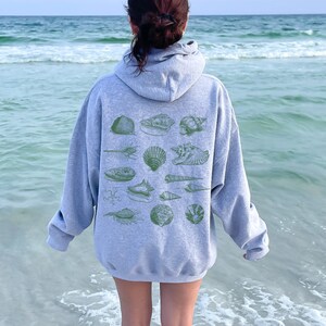 May include: A grey hooded sweatshirt with a design of various seashells in green on the back. The sweatshirt is being worn by a person standing on a beach.