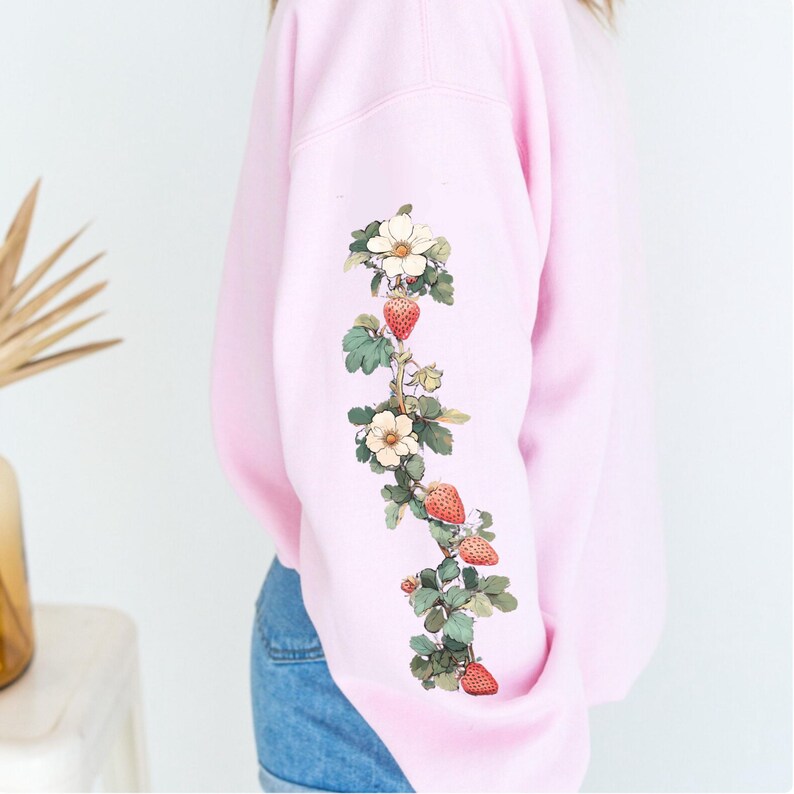 Peut inclure: Sweat-shirt rose avec un motif floral compos&eacute; de fleurs blanches et de fraises rouges sur la manche.