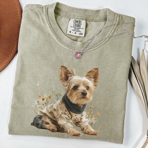 May include: A sage green Comfort Colors t-shirt featuring a Yorkshire Terrier wearing a floral sweater and a dark green scarf. A silver necklace with a pink pendant rests on the shirt. The shirt has a small tag that reads "S - P - CH".