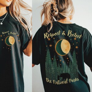 Op de afbeelding: Donkergrijs t-shirt met een grafisch ontwerp met een halve maan, sterren en dennenbomen. De tekst "Respect & Protect" staat boven de maan en "the National Parks" eronder. Een beer en welp staan in het bos.