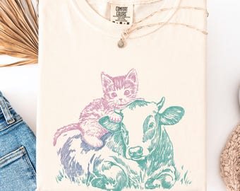 May include: Cream-colored t-shirt featuring a graphic of a kitten riding a cow, with the text "COMFORT COLORS" on the tag. The kitten is pink and purple, and the cow is teal and blue. A silver necklace with a small pendant is also visible.