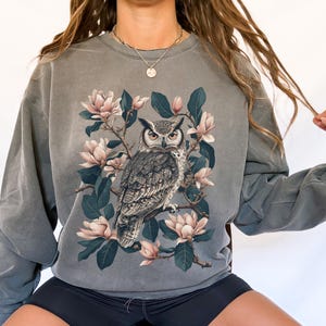 May include: A gray sweatshirt with a vintage-style print of an owl perched on a branch with magnolia blossoms. The owl is brown and gray with yellow eyes. The flowers are white and pink.