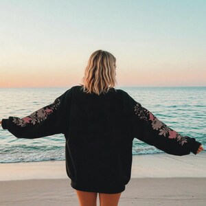 May include: Black sweatshirt with floral embroidery on the sleeves. The design features pink and beige flowers and leaves. The person is standing on a sandy beach with the ocean in the background.