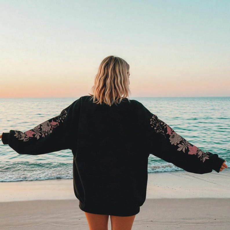 May include: Black sweatshirt with floral embroidery on the sleeves. The design features pink and beige flowers and leaves. The person is standing on a sandy beach with the ocean in the background.