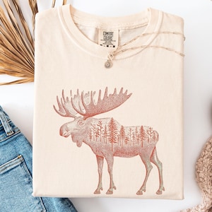 May include: Cream-colored t-shirt featuring a detailed illustration of a moose with a forest silhouette within its form. The shirt has a crew neck and short sleeves. The brand label "Comfort Colors" is visible on the upper left. A delicate necklace with a pendant rests on the shirt.