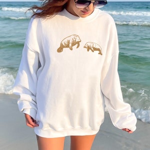 May include: White sweatshirt with a gold graphic of two manatees, one larger than the other, on the front. The sweatshirt is being worn by a person standing on a beach.