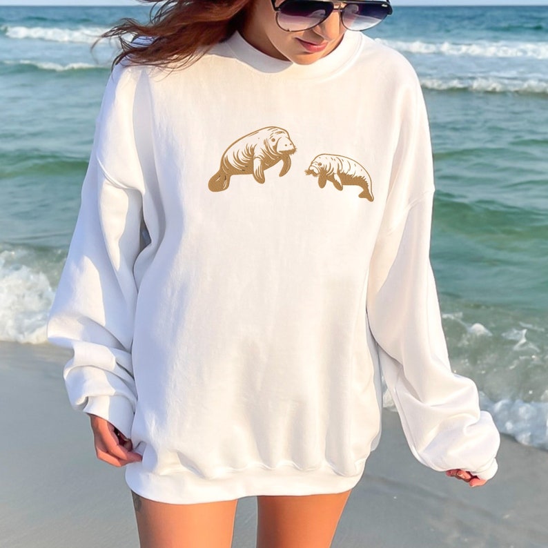 May include: White sweatshirt with a gold graphic of two manatees, one larger than the other, on the front. The sweatshirt is being worn by a person standing on a beach.