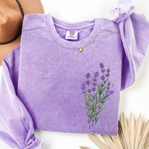 May include: A lavender-colored sweatshirt with a crew neck and long sleeves. The sweatshirt features an embroidered sprig of lavender flowers with green stems. A gold necklace with a pendant rests on the shirt. The brand tag is visible.