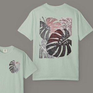 May include: Light green t-shirts with a monstera leaf design. The front of the shirt has a small leaf print, while the back features a larger, rectangular print of several leaves in shades of red, gray, and black.