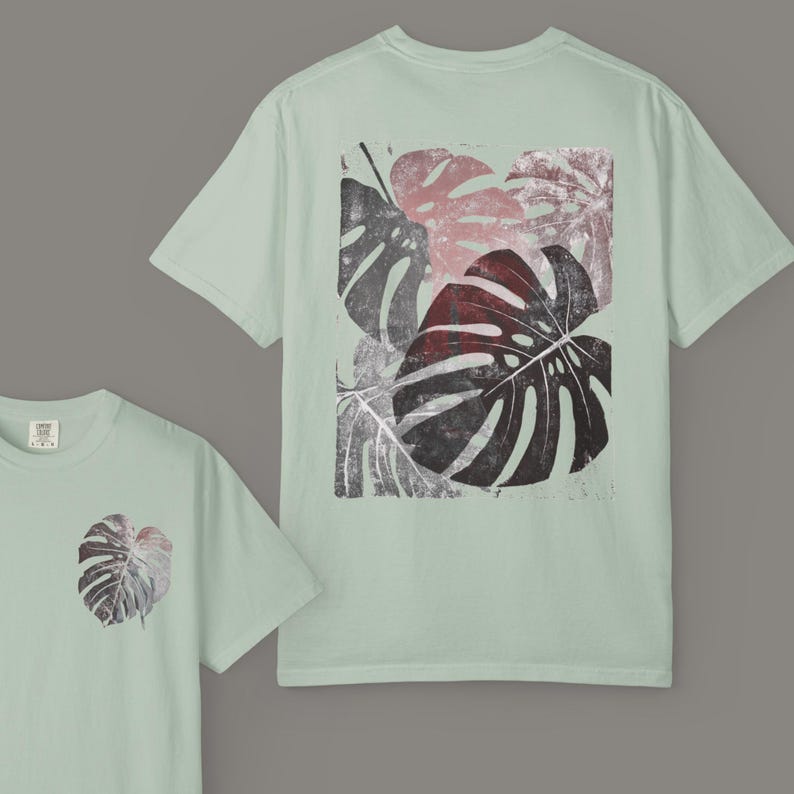 May include: Light green t-shirts with a monstera leaf design. The front of the shirt has a small leaf print, while the back features a larger, rectangular print of several leaves in shades of red, gray, and black.