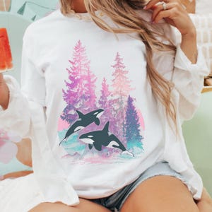 May include: White long-sleeve shirt featuring a graphic of two orcas swimming in front of a forest scene. The forest has pink, purple, and blue trees with a pink and orange sun in the background. The orcas are black and white.