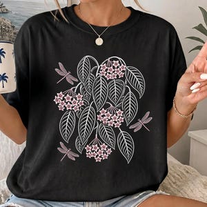 以下が含まれることがあります： 黒の半袖Tシャツ。中央に白い輪郭の葉と小さなピンクの星型花が集まったグラフィックプリントが施されている。ボタニカルイラストの周囲には3匹のピンクのトンボが描かれている。