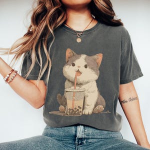 Op de afbeelding: Donkergrijs t-shirt met een cartoonkat die boba-thee drinkt. De kat is wit en grijs met een roze tong. De boba-thee zit in een doorzichtige beker. Het shirt heeft een relaxte pasvorm.