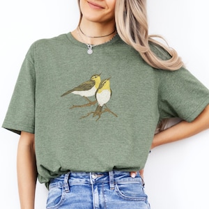 Op de afbeelding: Groen t-shirt met een geborduurd ontwerp van twee gele en bruine vogels op een tak. De vogels hebben gedetailleerde vleugels en staan tegen een gedempte groene achtergrond. Het shirt is gemaakt van een zacht materiaal.