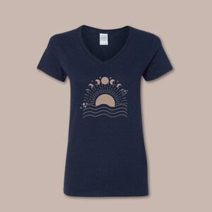 May include: Navy blue V-neck t-shirt featuring a celestial design. The graphic includes a sun, moon phases, stars, and wavy lines. The design is in a light brown color. The shirt is made of a soft material.