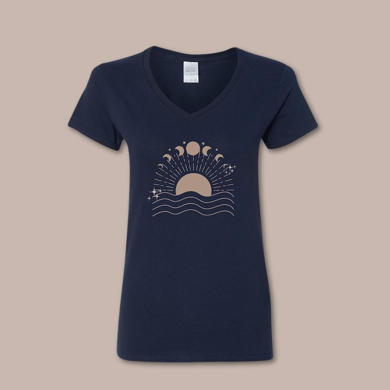 May include: Navy blue V-neck t-shirt featuring a celestial design. The graphic includes a sun, moon phases, stars, and wavy lines. The design is in a light brown color. The shirt is made of a soft material.