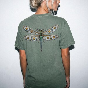 May include: Olive green t-shirt featuring a detailed dragonfly design on the back. The dragonfly's wings are adorned with white daisies and yellow centers. The insect's body is a mix of green, blue, and brown hues.